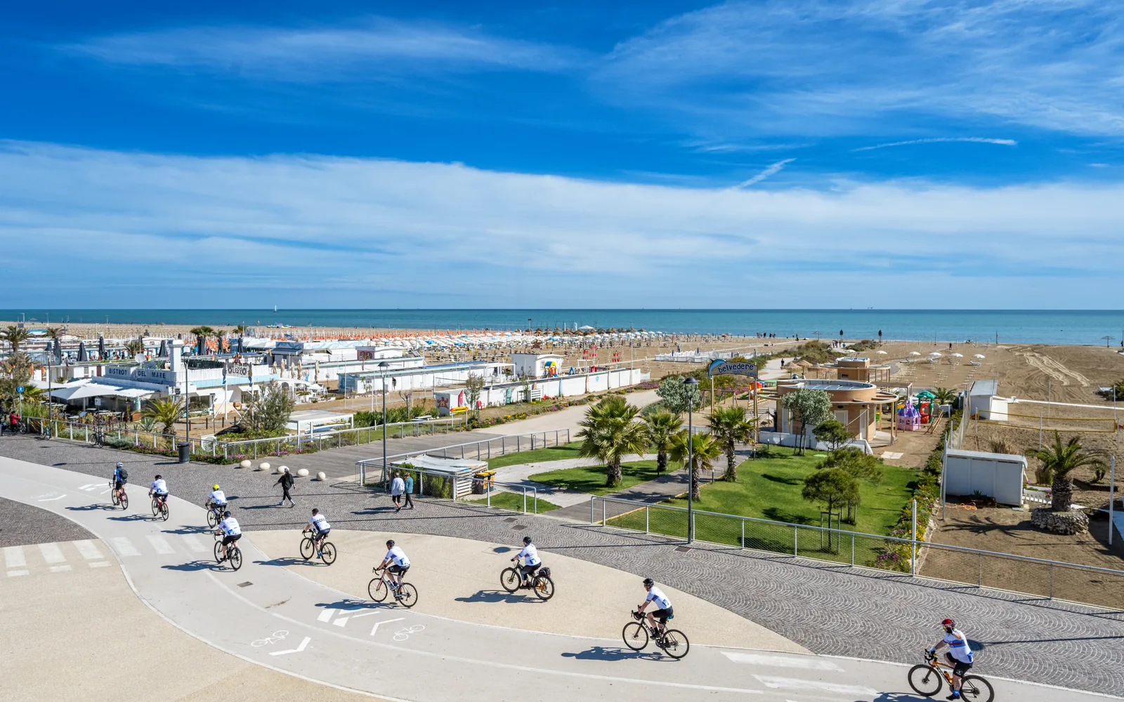 Ciclisti sulla pista ciclabile del lungomare verso Rimini, con spiagge e cabine sullo sfondo